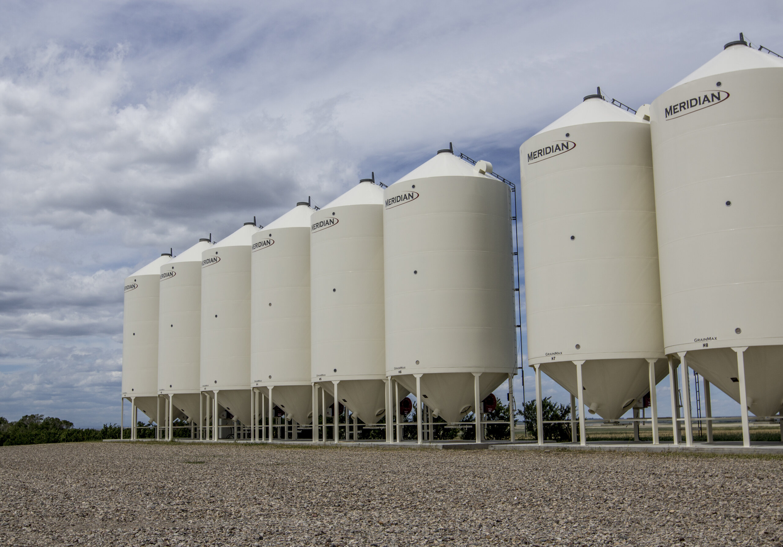 a row of ivory hopper bottom smoothwall grain bins against an overcast sky in a yard with gravel in front