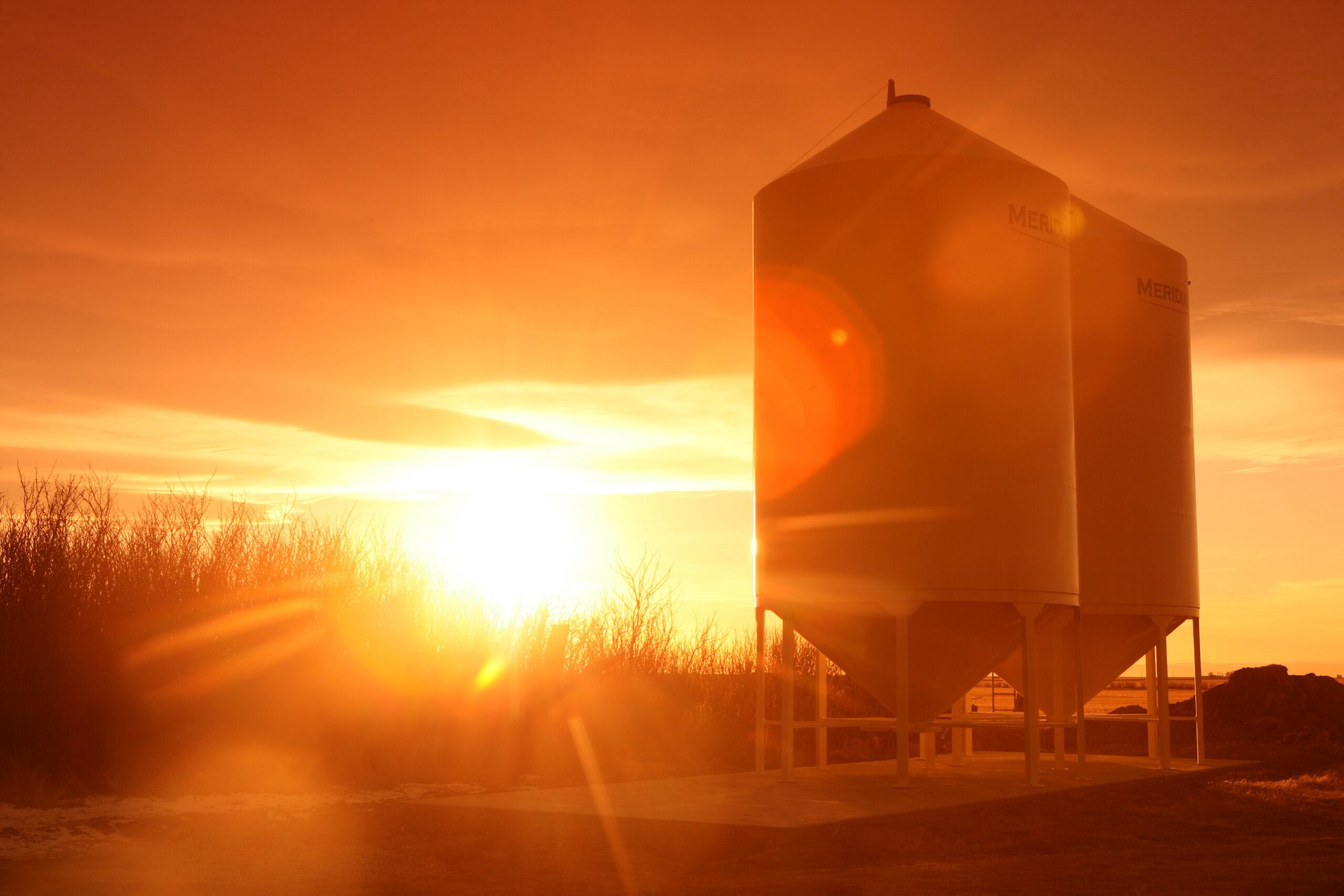 2 hopper bottom smoothwall grain bins silhouetted by an orange sunset