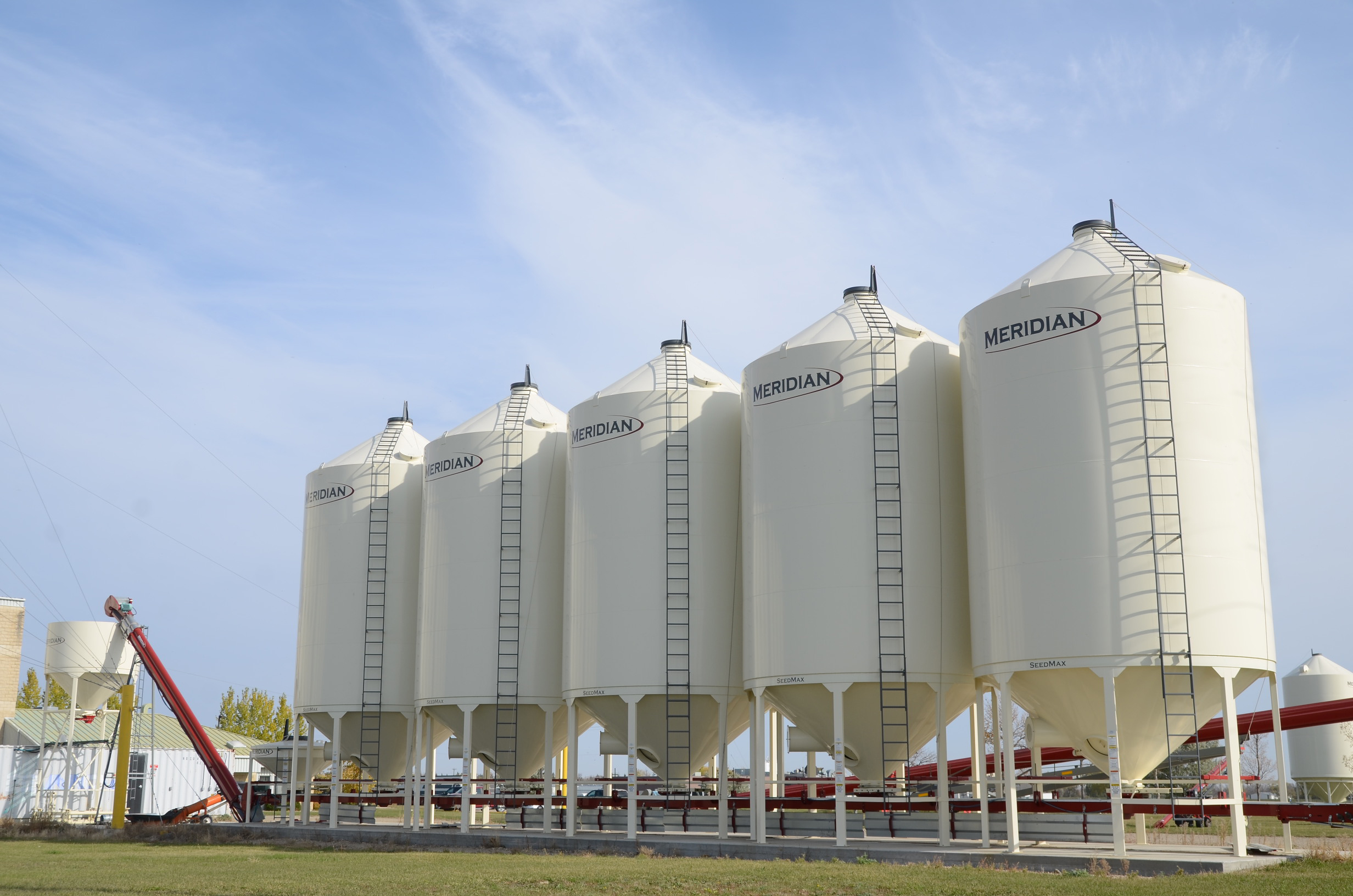 a row of ivory coloured seed bopper bottom smoothwall bins with a red underbin conveyor against a blue sky